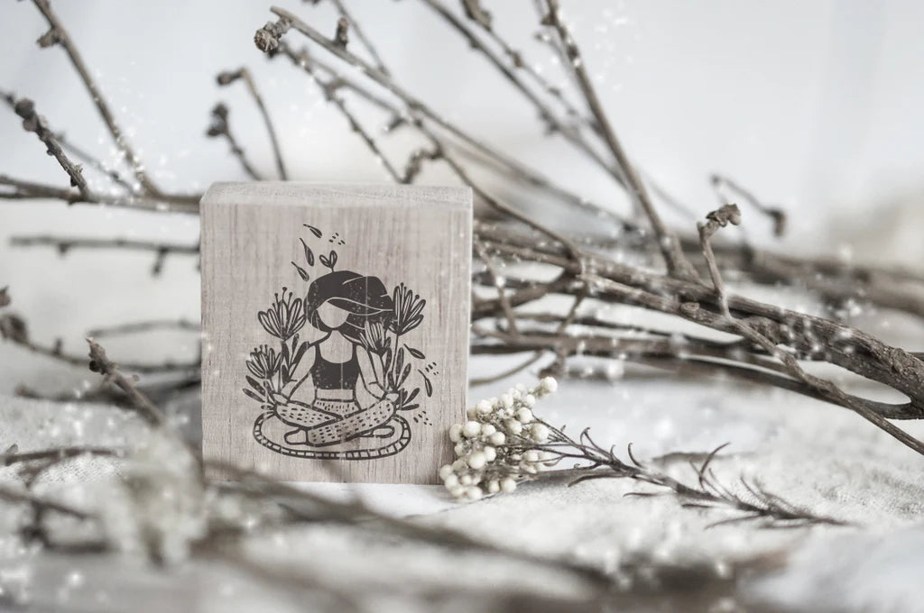 Wooden stamp with a design of a person meditating among plants, placed on a surface with branches and flowers.