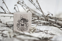 Wooden stamp with a design of a person meditating among plants, placed on a surface with branches and flowers.