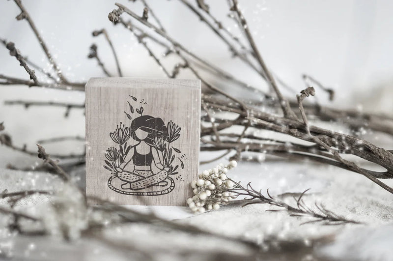 Wooden stamp with a design of a person meditating among plants, placed on a surface with branches and flowers.